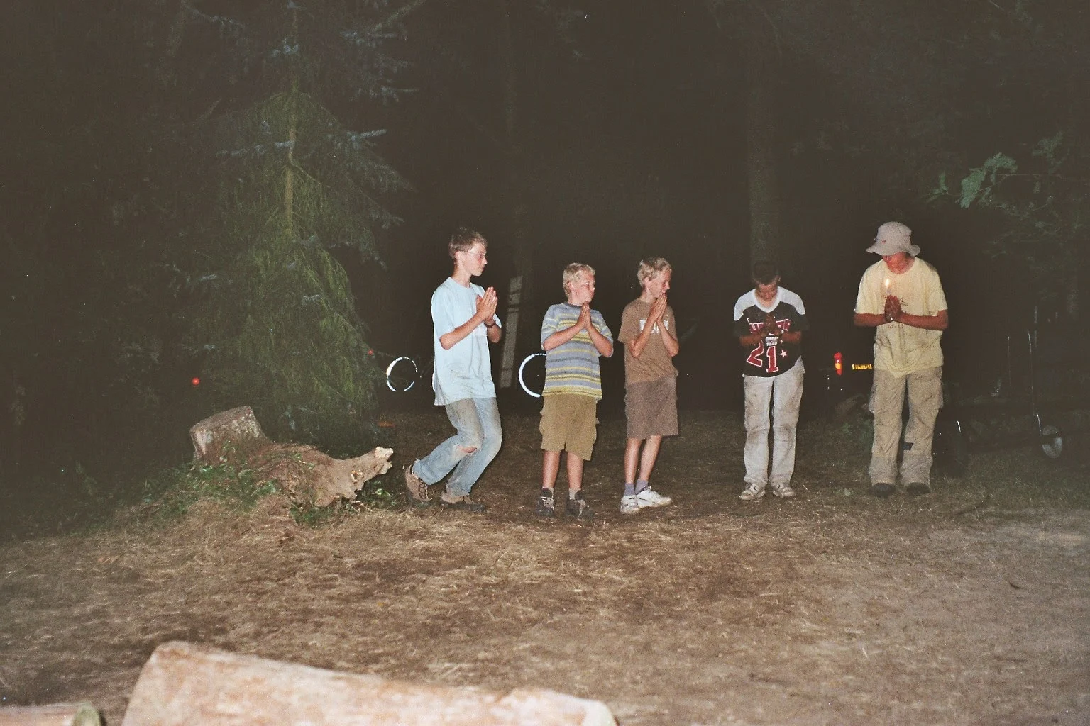 Kinderen en een leider staan 's avonds met gevouwen handen op een open plek in het bos